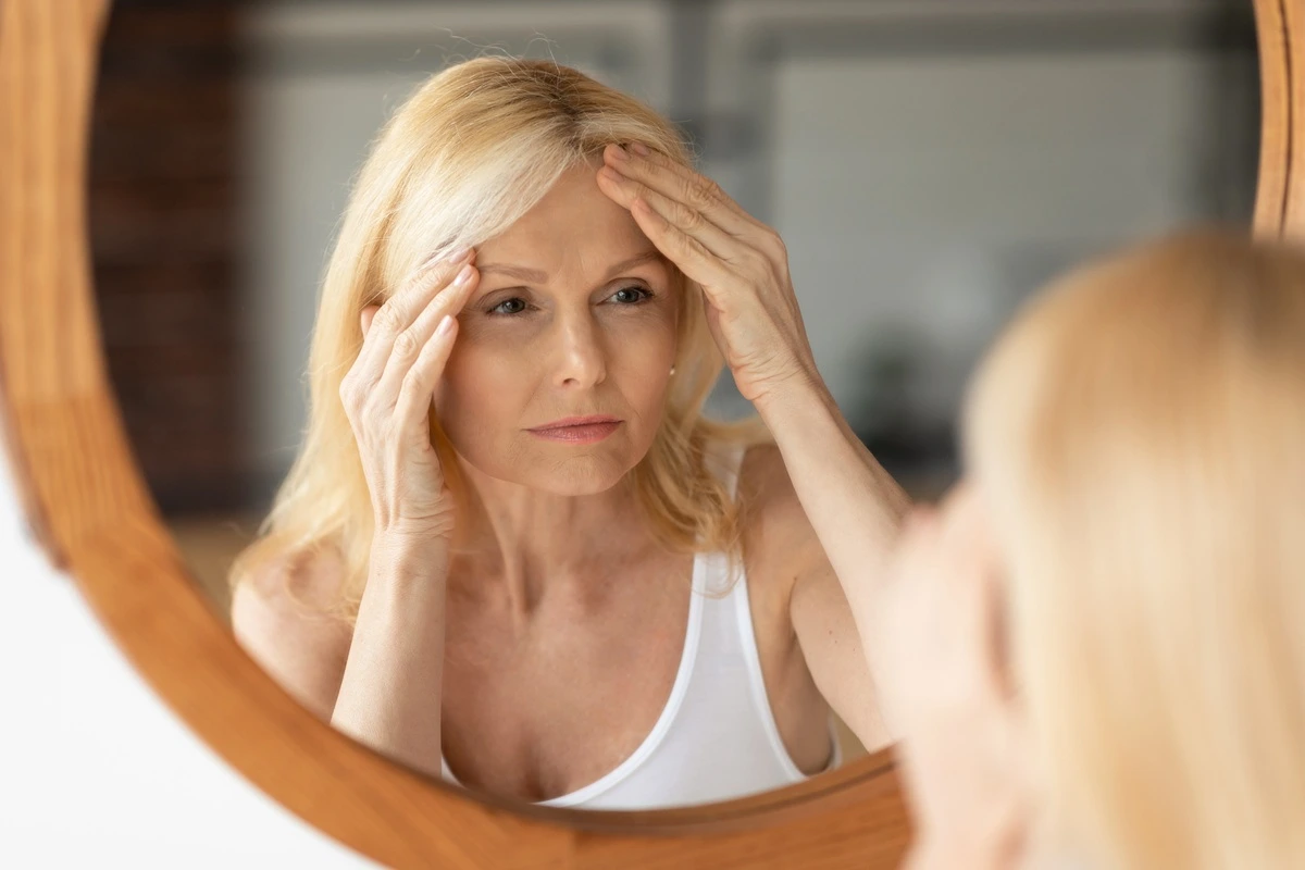 Mature blonde woman in a white tank top looking closely at her reflection in a round wooden mirror, touching her forehead to examine her skin and signs of aging.