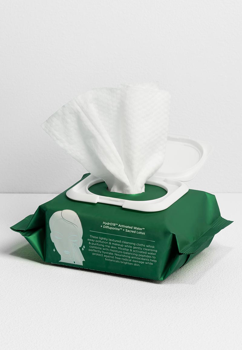 A dark green package of facial cleansing wipes with the white plastic lid open and a single white, textured cloth partially pulled out against a light green background.