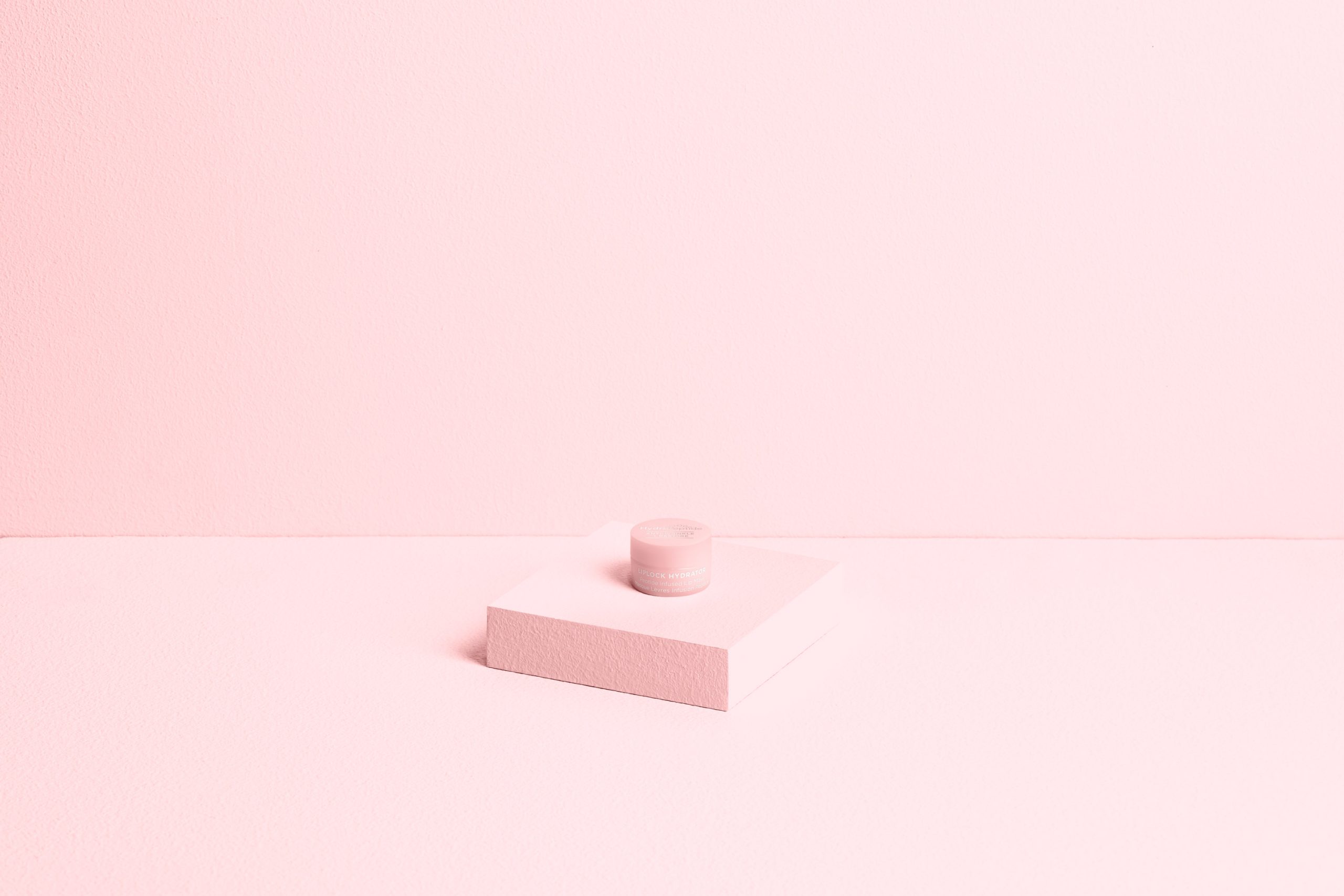 A small light pink jar on a pink square block against a light pink background.