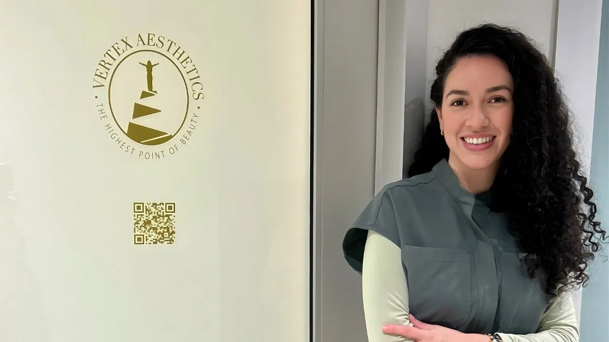 A smiling woman with long dark curly hair wearing grey-green scrubs and a light long-sleeve undershirt stands beside a wall featuring the gold circular logo and branding for "VERTEX AESTHETICS - THE HIGHEST POINT OF BEAUTY" along with a QR code.